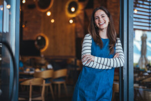 A joyful woman in a striped shirt and denim apron stands with arms crossed at a welcoming coffee house